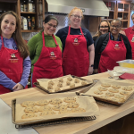 Six people wearing aprons stand in front of cookie sheets in a kitchen at the Ronald McDonald House.