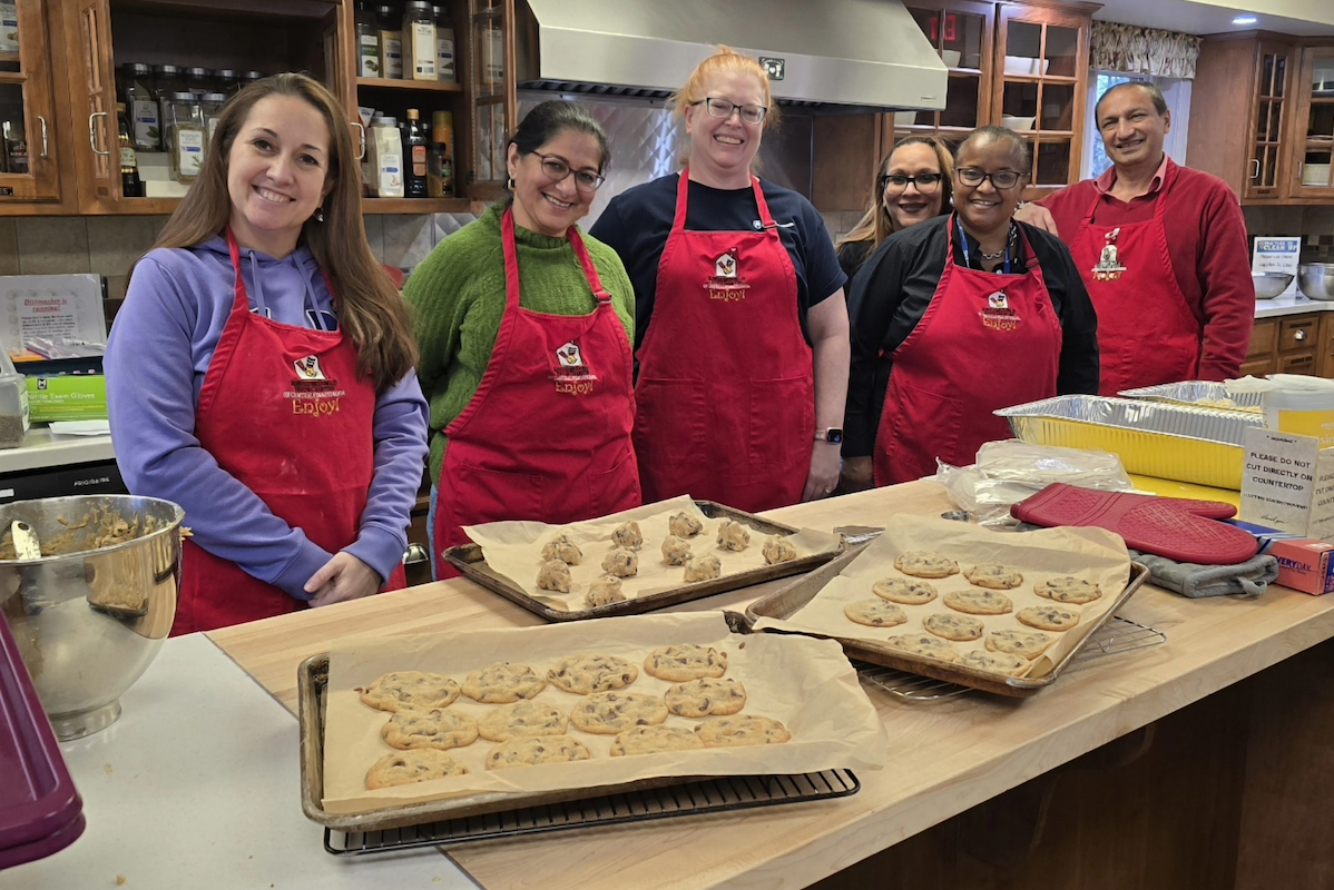 Six people wearing aprons stand in front of cookie sheets in a kitchen at the Ronald McDonald House.