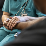 A health care professional in scrubs sits next to a patient, holding their hand.