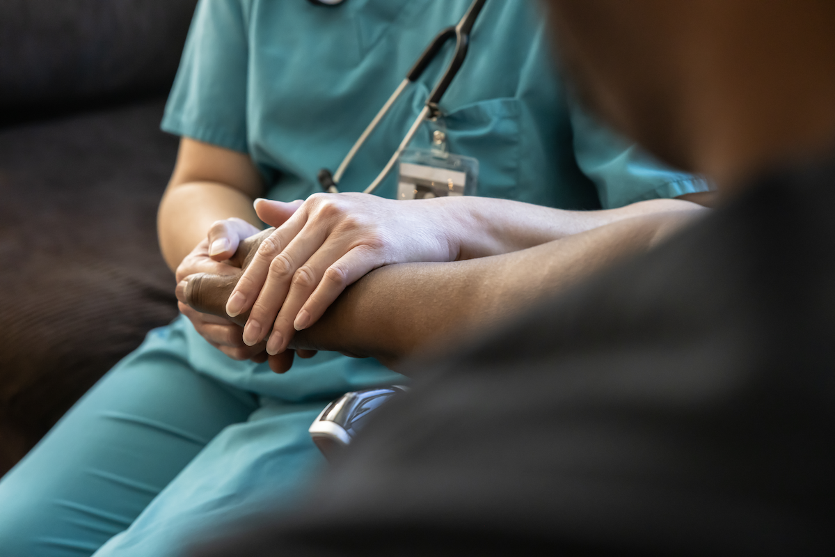 A health care professional in scrubs sits next to a patient, holding their hand.
