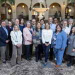 A group of approximately 23 people in business casual attire pose for a group photo in a hotel lobby. A fountain is in the near background.