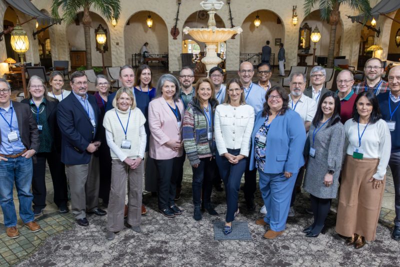 A group of approximately 23 people in business casual attire pose for a group photo in a hotel lobby. A fountain is in the near background.