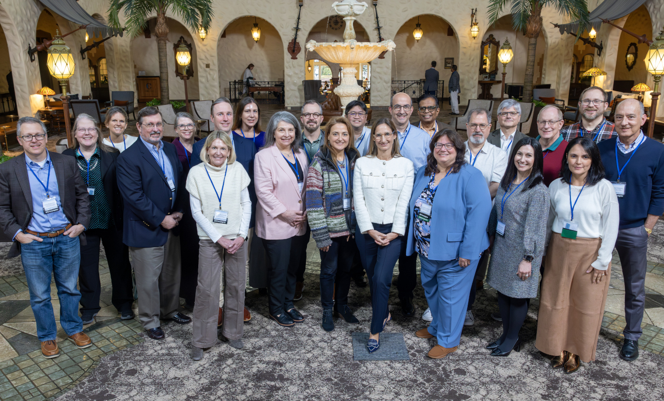 A group of approximately 23 people in business casual attire pose for a group photo in a hotel lobby. A fountain is in the near background.
