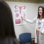 Dr. Christina DeAngelis, wearing a white lab coat and name badge, points to a chart about the female reproductive. A woman patient is seen from behind.
