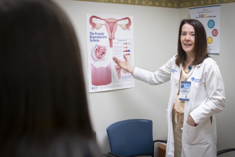 Dr. Christina DeAngelis, wearing a white lab coat and name badge, points to a chart about the female reproductive. A woman patient is seen from behind.