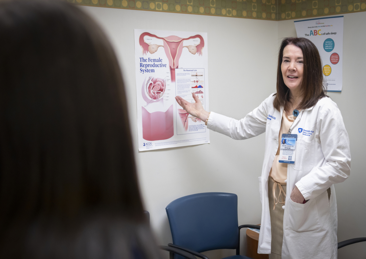 Dr. Christina DeAngelis, wearing a white lab coat and name badge, points to a chart about the female reproductive. A woman patient is seen from behind.