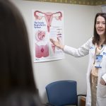 Dr. Christina DeAngelis, wearing a white lab coat and name badge, points to a chart about the female reproductive. A woman patient is seen from behind.