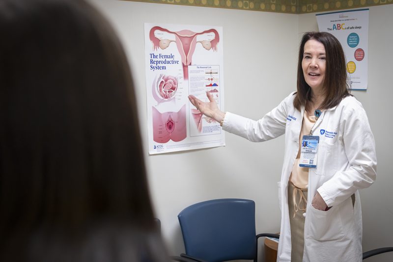 Dr. Christina DeAngelis, wearing a white lab coat and name badge, points to a chart about the female reproductive. A woman patient is seen from behind.