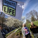 A man and a woman stand in a grassy area looking up at a ‘Donate Life’ flag hoisted several feet into the air on a flagpole. In the background are a sidewalk, a large building, some trees and bushes, and a TV videographer.