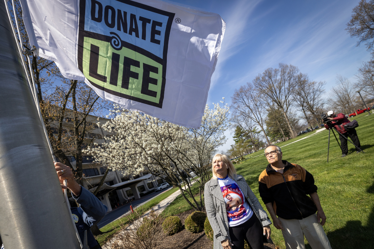 A man and a woman stand in a grassy area looking up at a ‘Donate Life’ flag hoisted several feet into the air on a flagpole. In the background are a sidewalk, a large building, some trees and bushes, and a TV videographer.