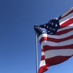 The American flag on a flying on a flagpole, a clear sky in the background.
