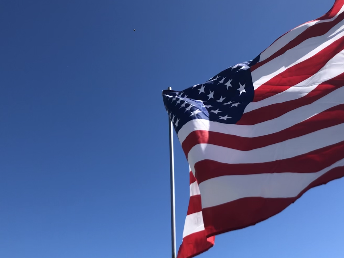 The American flag on a flying on a flagpole, a clear sky in the background.
