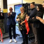 Six people smile and clap immediately after cutting a ribbon, as they stand in front of a vending machine. A window is also in the background.