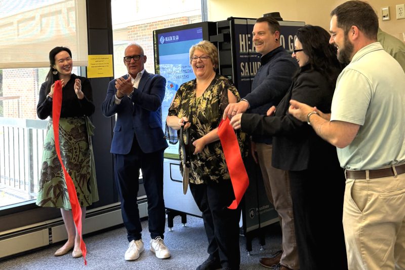 Six people smile and clap immediately after cutting a ribbon, as they stand in front of a vending machine. A window is also in the background.