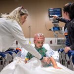 A young child wearing a monitoring cap sits on a hospital bed while two medical staff assist with care and adjust medical equipment.