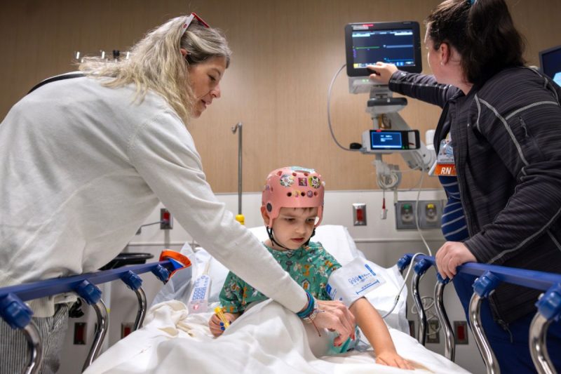 A young child wearing a monitoring cap sits on a hospital bed while two medical staff assist with care and adjust medical equipment.