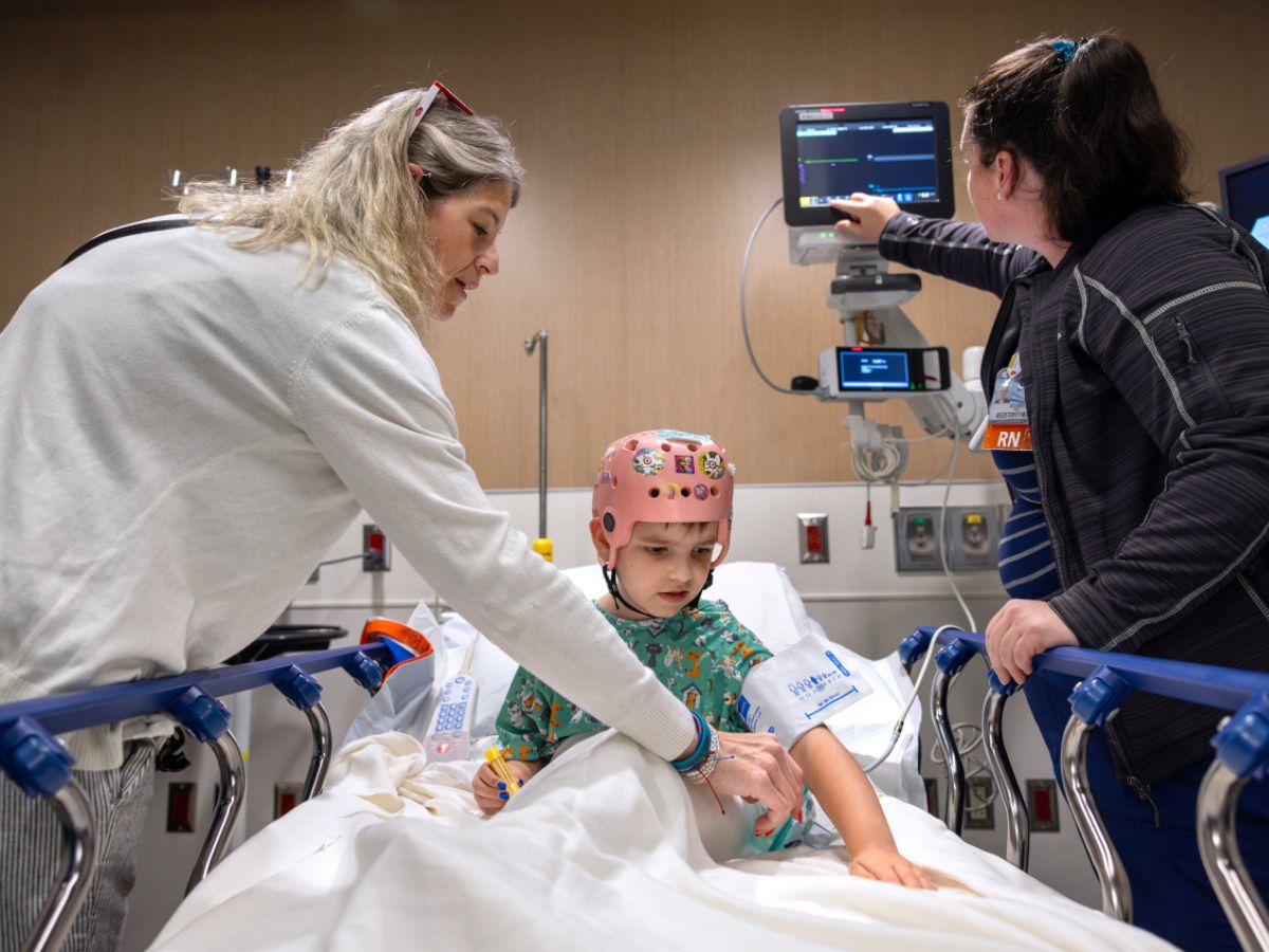 A young child wearing a monitoring cap sits on a hospital bed while two medical staff assist with care and adjust medical equipment.