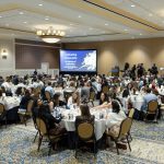 Students, faculty, staff and community partners sit at round banquet tables during the OHACE year-end celebration at Hershey Lodge, facing a stage with a screen displaying the words “Cultivating Community: An Evening of Celebration.”
