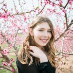 A teen girl in front of a flowering tree smiles at the camera. She has long hair and is touching her shoulder.