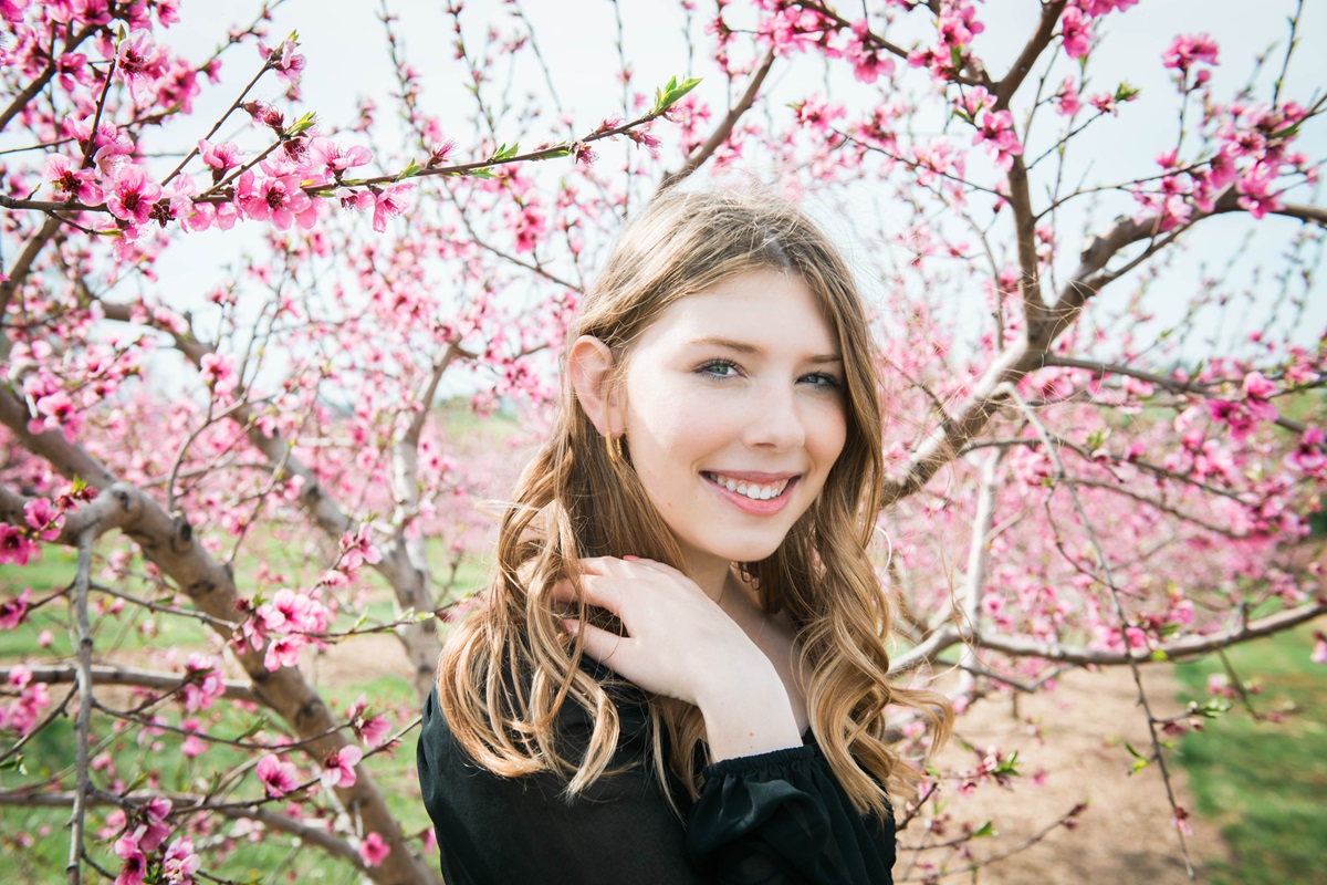 A teen girl in front of a flowering tree smiles at the camera. She has long hair and is touching her shoulder.