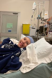 Keira Woods smiles as she lies in bed in a hospital room. She is covered with a blanket and has a tight cap on her head. Behind her on the right is an IV pole with two IV bags and other medical equipment. A door is behind her on the left.