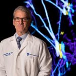Jeffrey Miller stands in his white doctor's coat with a digital dermatology display behind him.
