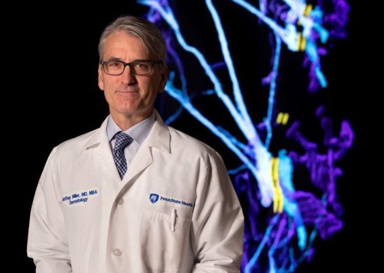 Jeffrey Miller stands in his white doctor's coat with a digital dermatology display behind him.