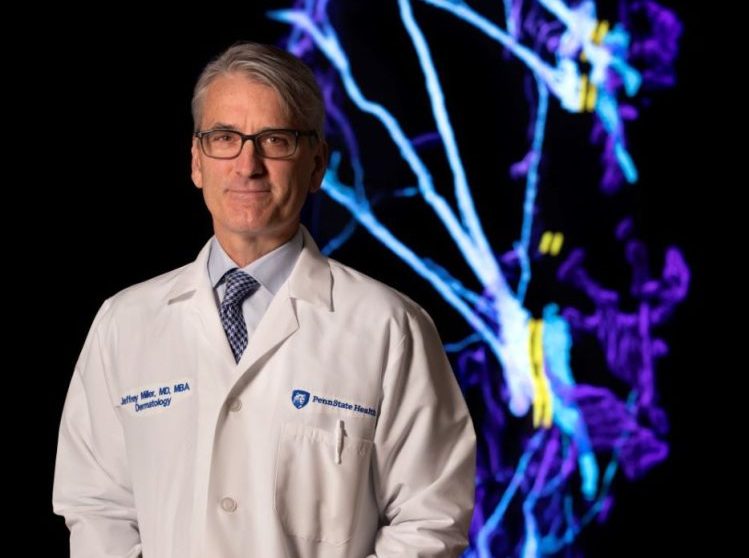 Jeffrey Miller stands in his white doctor's coat with a digital dermatology display behind him.