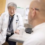 A physician wearing a white medical coat and a nametag is seated sideways in a clinic room, speaking with a patient who is in the foreground, slightly out of focus and looking away from the camera.
