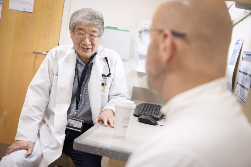 A physician wearing a white medical coat and a nametag is seated sideways in a clinic room, speaking with a patient who is in the foreground, slightly out of focus and looking away from the camera.