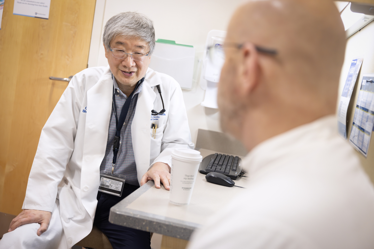 A physician wearing a white medical coat and a nametag is seated sideways in a clinic room, speaking with a patient who is in the foreground, slightly out of focus and looking away from the camera.