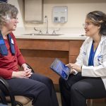 A woman patient sits across from a neurologist in a medical office. The patient has an adaptive deep brain stimulation device around her neck. The neurologist touches a tablet to adjust the device and smiles at the patient.