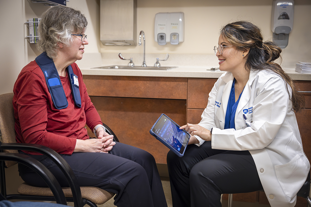 A woman patient sits across from a neurologist in a medical office. The patient has an adaptive deep brain stimulation device around her neck. The neurologist touches a tablet to adjust the device and smiles at the patient.