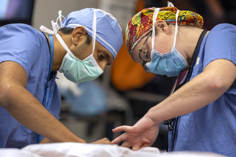 Two surgeons operate on a patient. The man on the left is wearing a face mask and scrubs. The woman on the right is wearing a colorful surgical cap, glasses, face mask and scrubs.