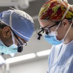 Two surgeons look down as they perform a procedure. The man on the left is wearing a face mask, scrubs, and specialized magnifying glasses called surgical loupes. The woman on the right is wearing a colorful surgical cap, glasses, surgical loupes face mask and scrubs.