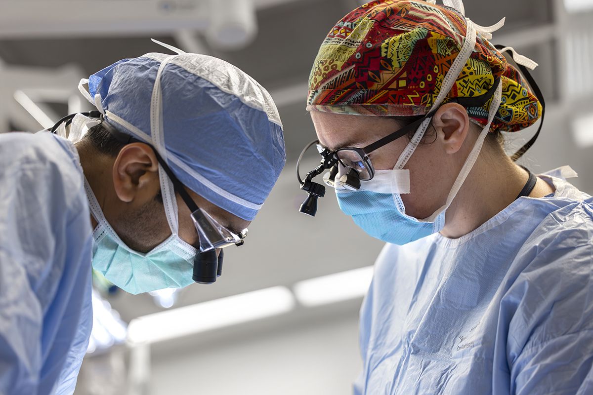 Two surgeons look down as they perform a procedure. The man on the left is wearing a face mask, scrubs, and specialized magnifying glasses called surgical loupes. The woman on the right is wearing a colorful surgical cap, glasses, surgical loupes face mask and scrubs.