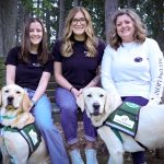Three women sit on a wooden bench outdoors in a wooded area, smiling at the camera, each accompanied by a Golden Labrador retriever wearing a green “Canine Assistants” service vest.
