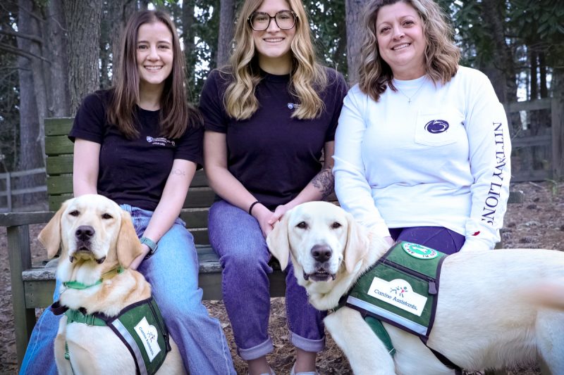 Three women sit on a wooden bench outdoors in a wooded area, smiling at the camera, each accompanied by a Golden Labrador retriever wearing a green “Canine Assistants” service vest.