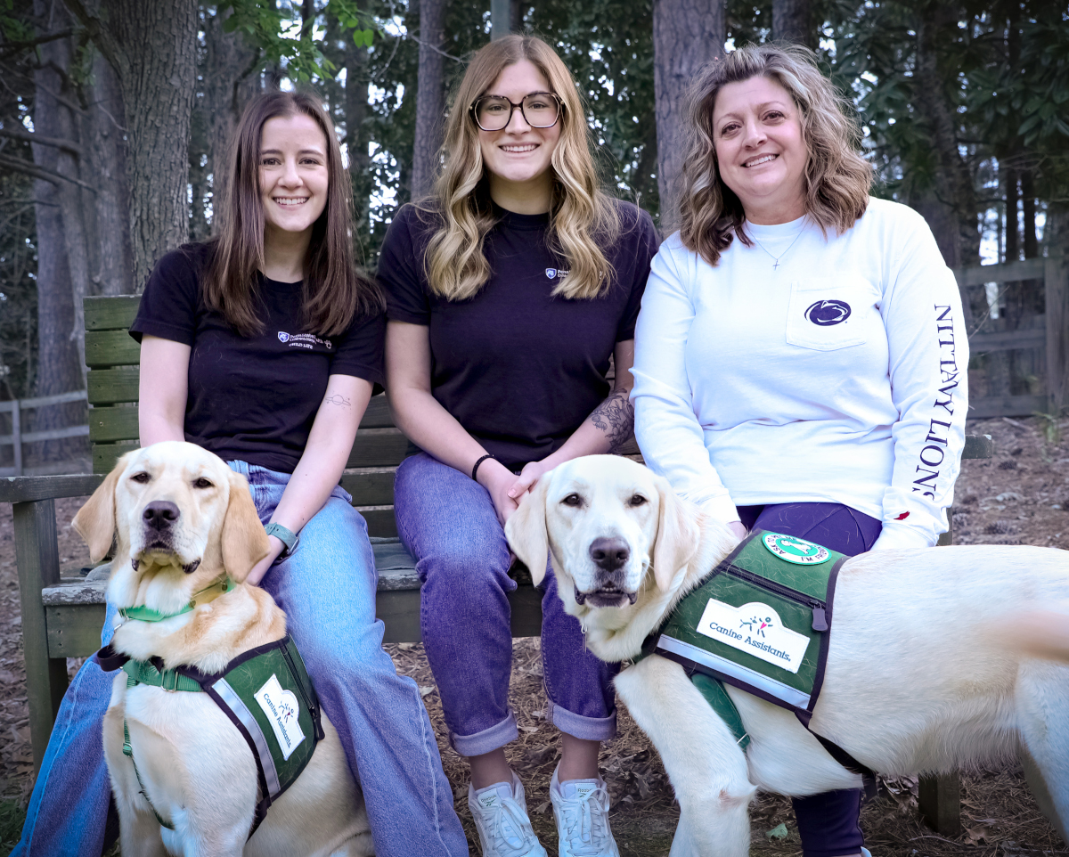 Three women sit on a wooden bench outdoors in a wooded area, smiling at the camera, each accompanied by a Golden Labrador retriever wearing a green “Canine Assistants” service vest.