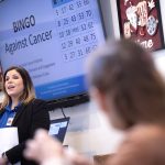A woman stands in front of a screen with the words “BINGO Against Cancer” on it and a bingo card. She puts two fingers in the air. A woman, out of focus on the right, watches her.