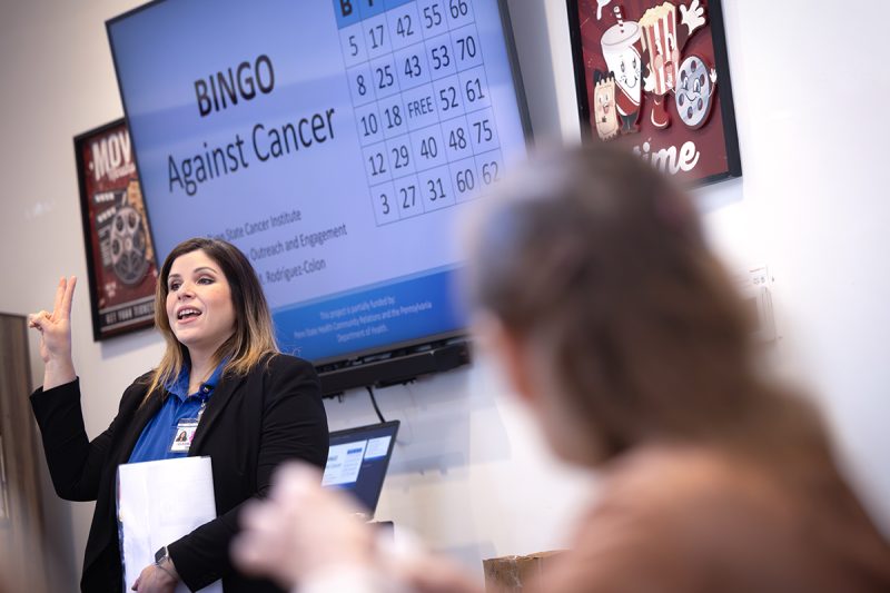 A woman stands in front of a screen with the words “BINGO Against Cancer” on it and a bingo card. She puts two fingers in the air. A woman, out of focus on the right, watches her.