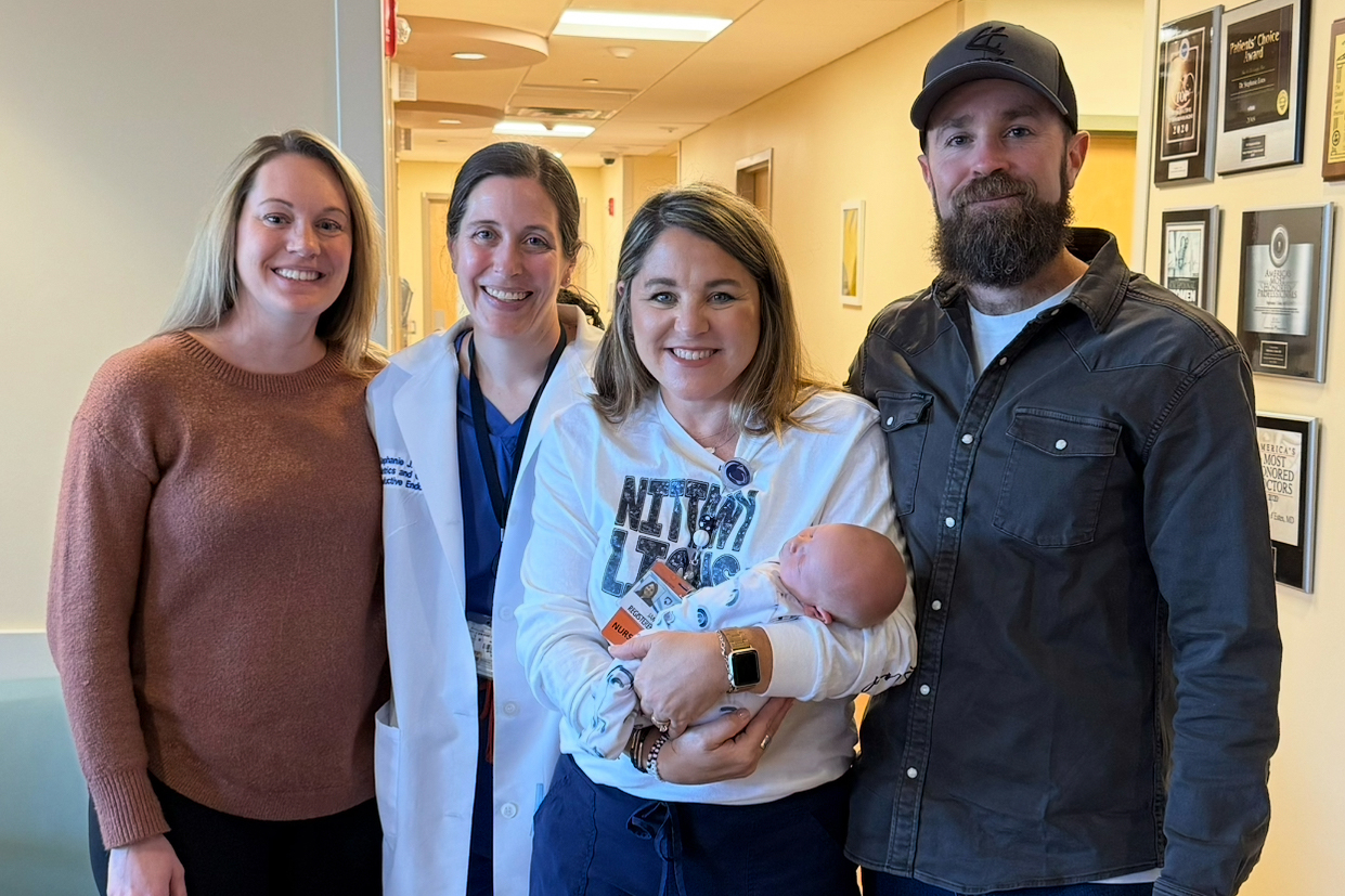 A group of four adults stands together with the woman in the center holding a baby. Pictures hang on the wall in the hallway behind them.