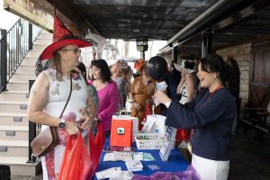 A woman wearing a witch’s hat talks to a smiling woman at a table filled with cards and brochures. Behind them is a group of women.