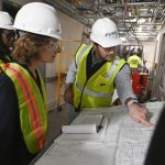 A man in a hard hat points to blueprints laid out on a table while two women in hard hats look on. They’re standing in a hallway that’s under construction.