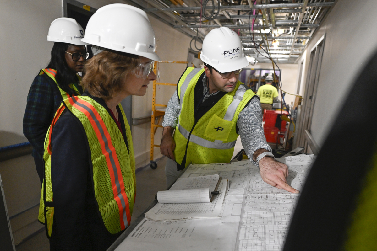 A man in a hard hat points to blueprints laid out on a table while two women in hard hats look on. They’re standing in a hallway that’s under construction.