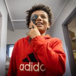 A boy holds an occluder over his right eye and smiles as he looks at a vision chart. He is wearing a sweatshirt with an Adidas logo on it.