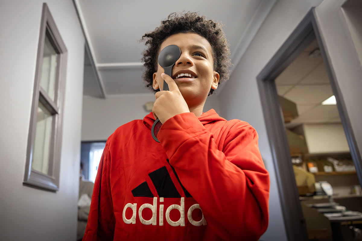 A boy holds an occluder over his right eye and smiles as he looks at a vision chart. He is wearing a sweatshirt with an Adidas logo on it.