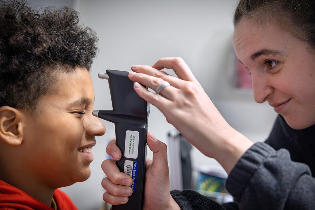 A boy squints as he gets an eye pressure test from an ophthalmic technician. The woman technician on the right smiles as she holds a tonometer in front of his eye.