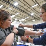 A woman holds a blood pressure cuff on her arm as a nurse on the right adjusts it. They are sitting at a table in a large warehouse room with a high ceiling and bright fluorescent lights.