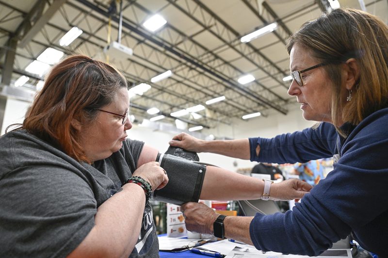 A woman holds a blood pressure cuff on her arm as a nurse on the right adjusts it. They are sitting at a table in a large warehouse room with a high ceiling and bright fluorescent lights.
